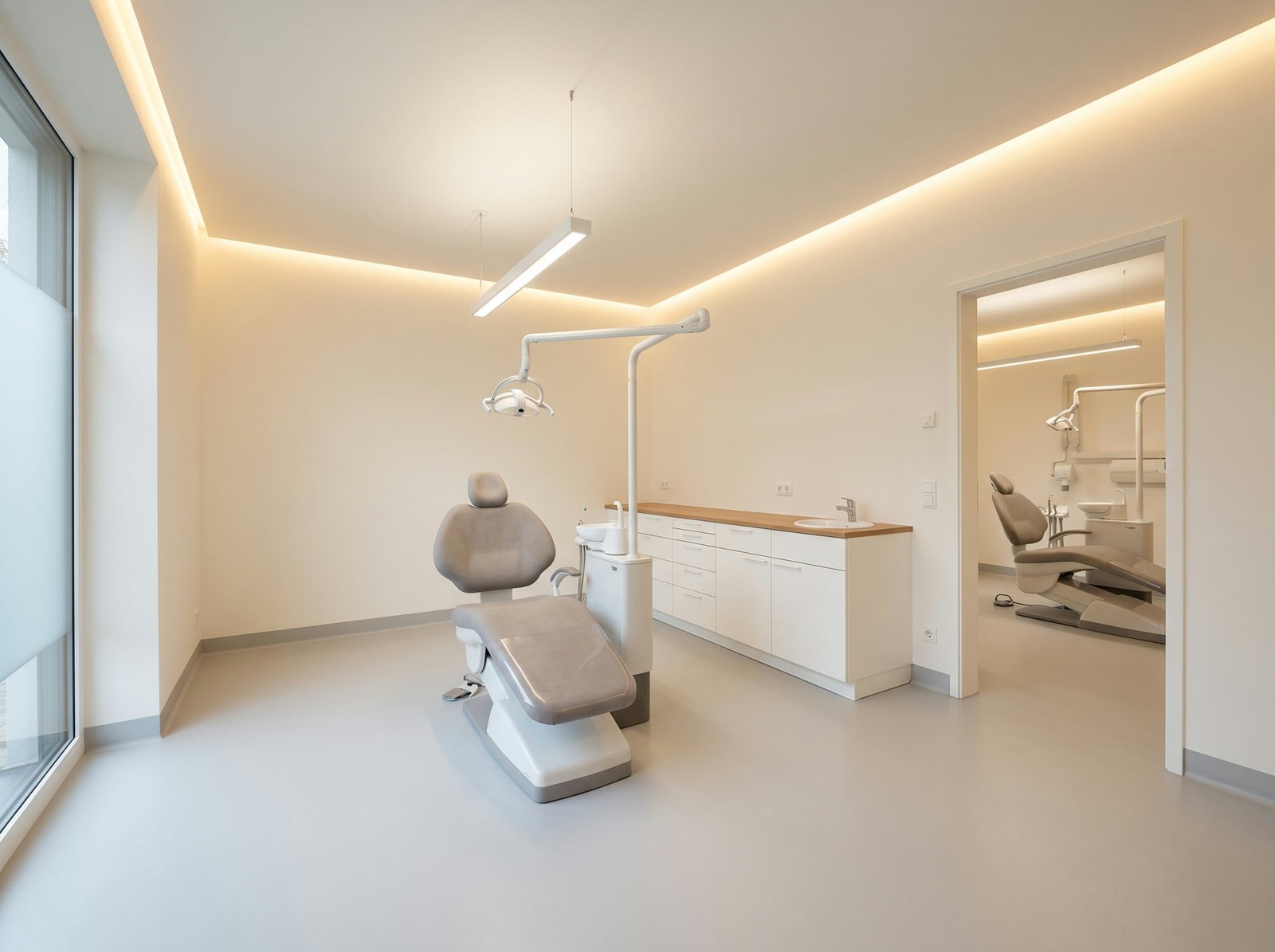 Blick durch zwei Behandlungszimmer einer Zahnarztpraxis mit Behandlungseinheiten, Unterschrankzeile mit Eichenarbeitsplatte und indirekter Beleuchtung
